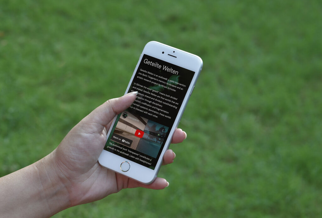 A hand holding a smartphone, against a grassy background. The screen of the phone shows the home page of the Geteilte Welten website.